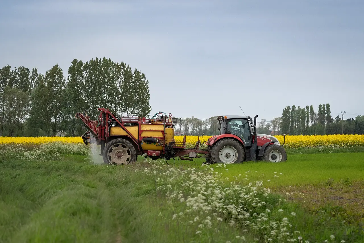 A vibrant scene of precision agriculture: a red tractor pulls a large yellow and red crop sprayer actively applying a fine mist of liquid. The machinery works amidst foreground weeds with white flowers, set against a vast, bright yellow rapeseed field, distant trees, and a clear blue sky.
