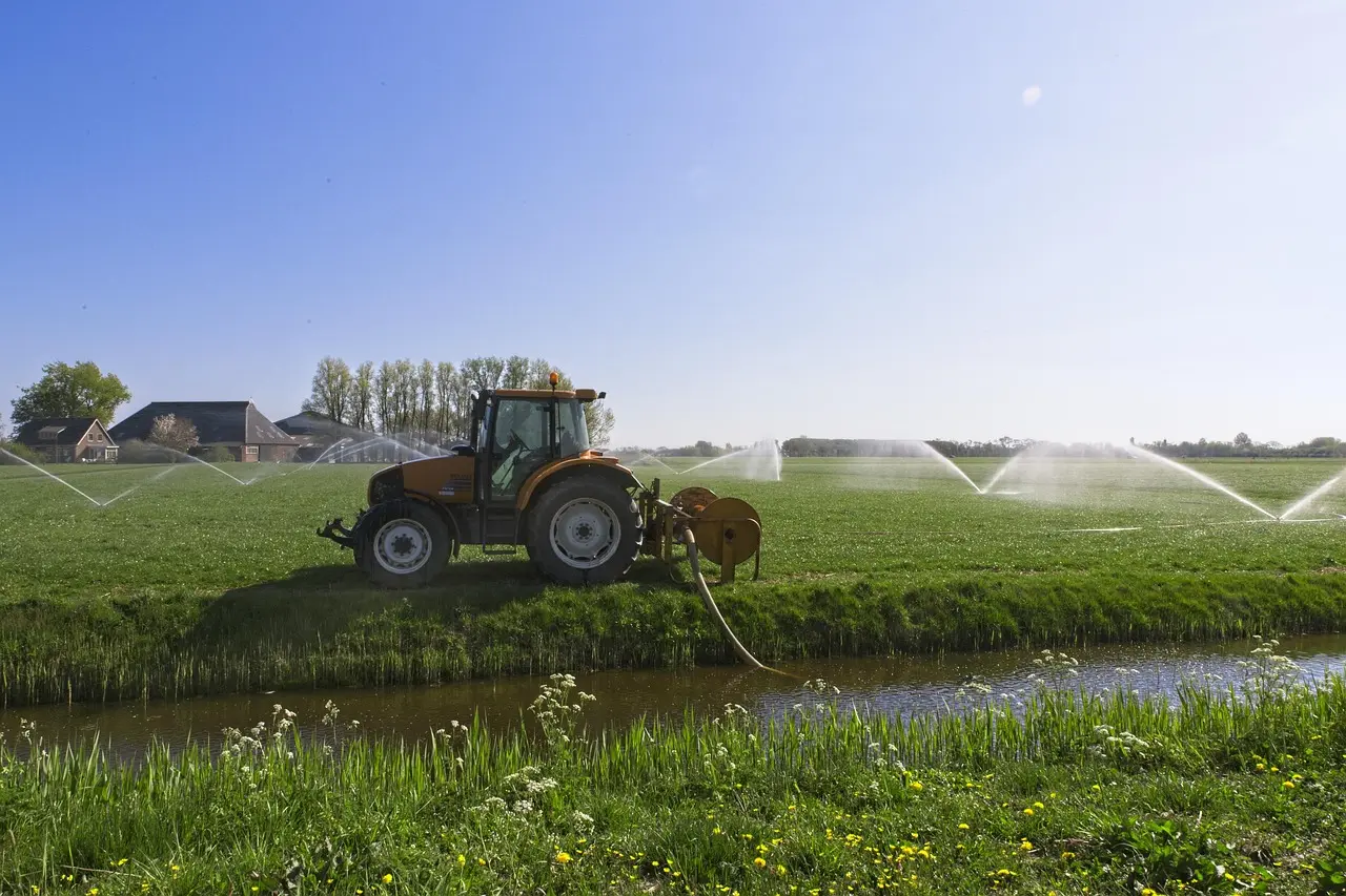 An orange tractor is parked in a vast green field, towing a large irrigation system with multiple sprinklers that are actively spraying arcs of water mist into the distance. White wildflowers dot the grass, and a narrow irrigation channel runs in the foreground. The scene is set under a clear blue sky with distant trees and a brown-roofed house, conveying a peaceful, productive rural atmosphere.