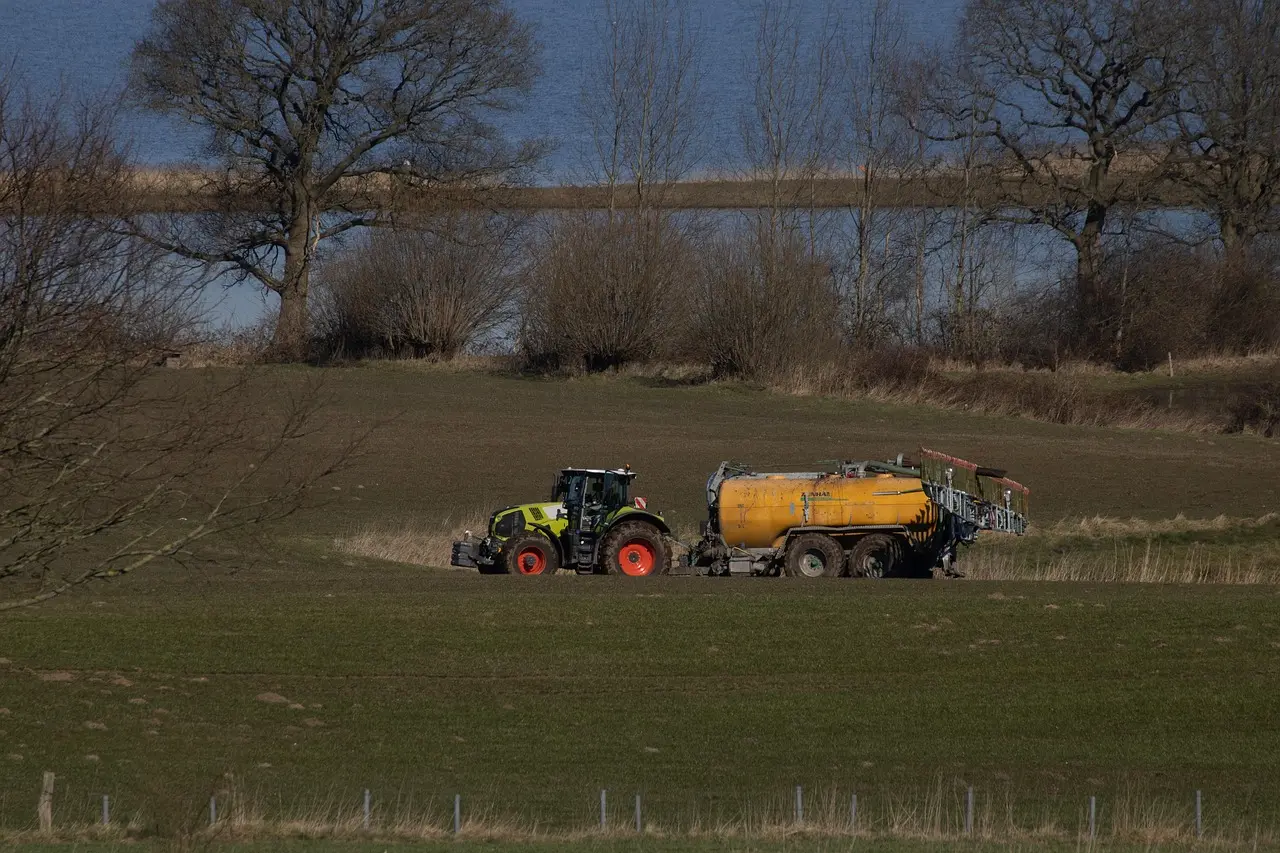 A bright green tractor with red wheel hubs tows a large yellow agricultural sprayer or fertilizer tank across a field. The scene, set against a backdrop of leafless trees, a dense shrub line, and a distant body of water, is illuminated by the low-angle light of early morning or late afternoon, casting long shadows and conveying a peaceful, productive rural atmosphere.