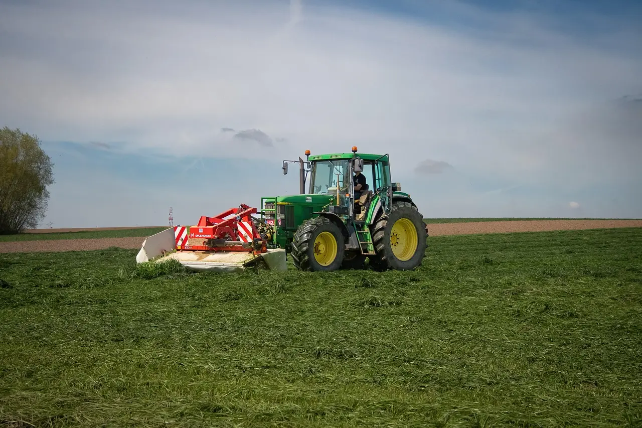 A green tractor with yellow tires operates in a vast green field, pulling a red agricultural implement for tilling or harvesting. An operator is visible in the cab. The scene, under a partly cloudy sky, showcases modern machinery at work in an expansive farming landscape.