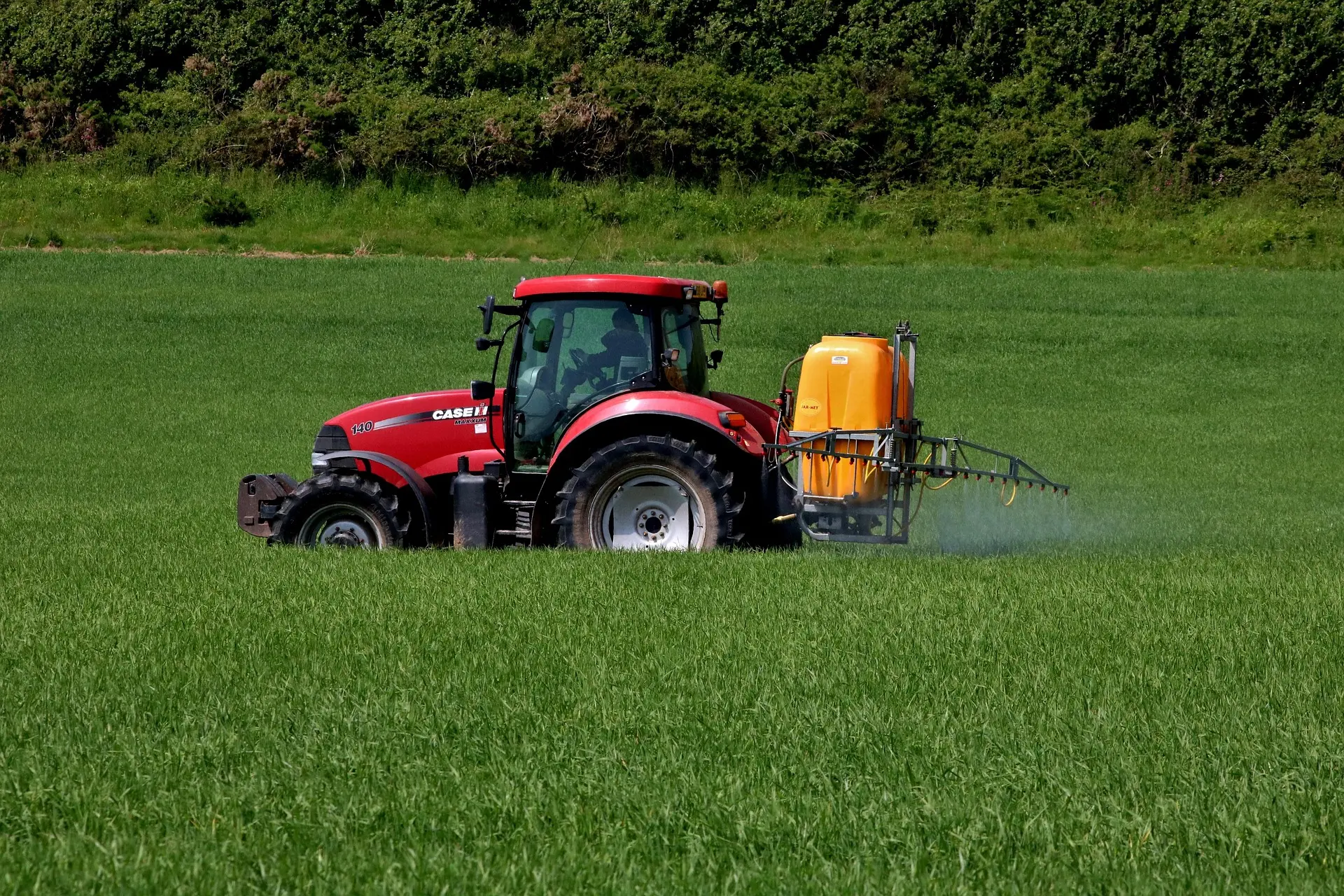 A red CASE IH tractor, model 140, with a yellow front frame, tows a yellow sprayer applying mist to a vibrant green field. Lush trees and shrubs are in the background under a clear sky.
