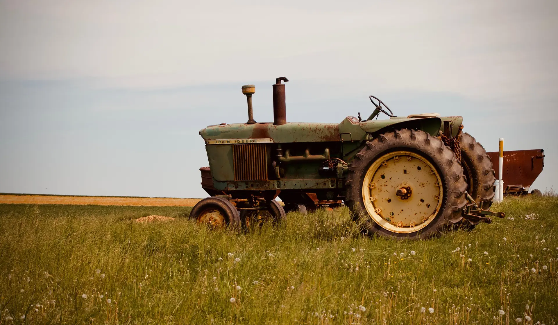 An old, weathered green John Deere tractor with heavy rust on its body and large rear tires sits at an angle in an overgrown field, with white dandelion seeds in the foreground under a cloudy sky.