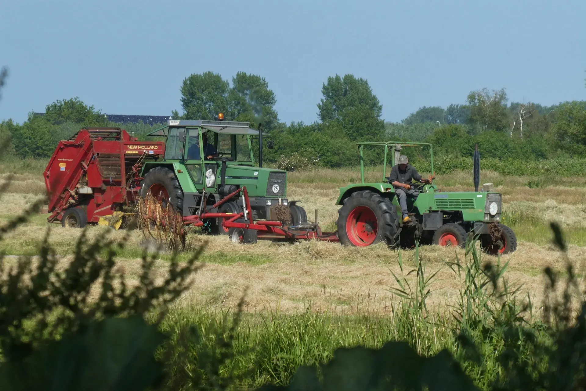 Two green tractors work together in a hayfield: a smaller one with an operator tows a large red New Holland baler, while a larger tractor follows behind.