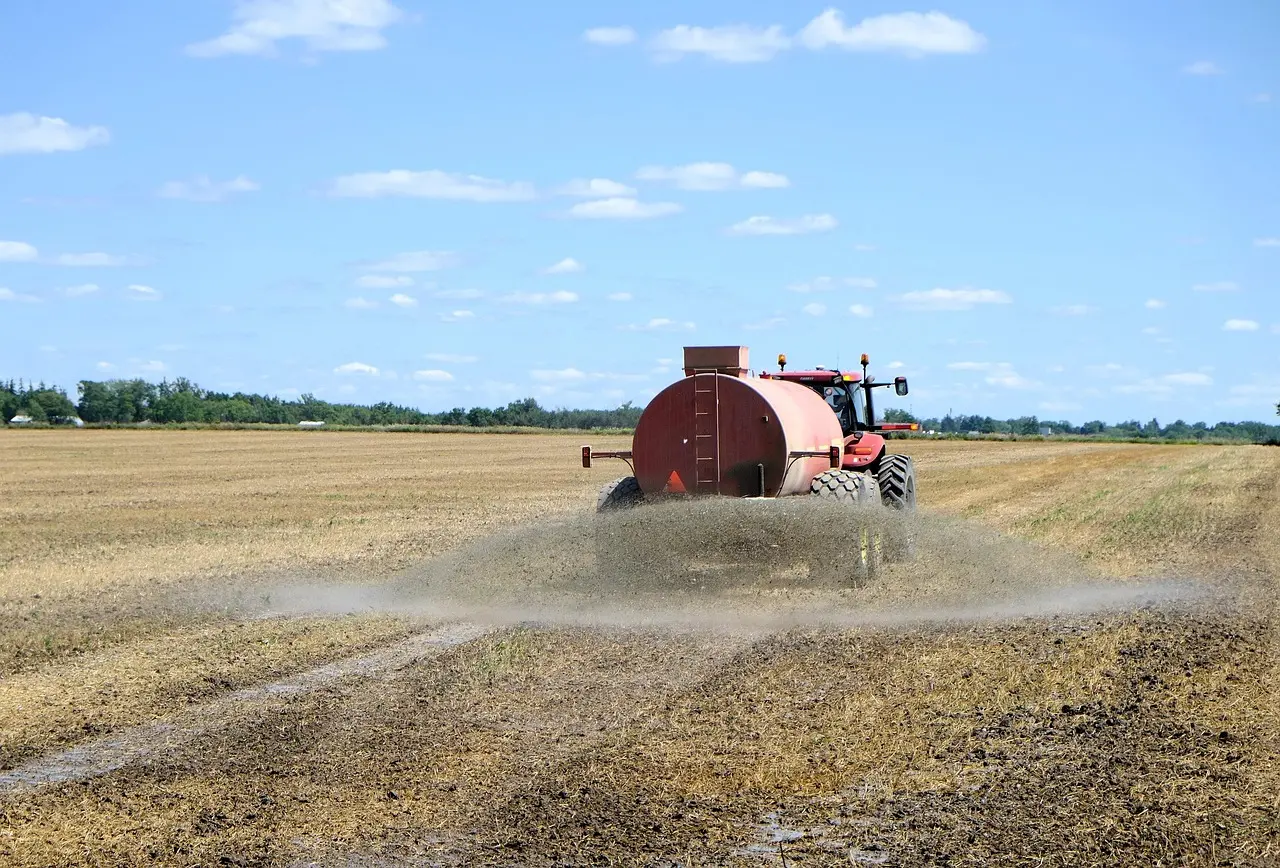 A red tractor towing a large cylindrical tank kicks up dust as it moves across a vast, open field under a clear blue sky with white clouds, with distant trees and buildings in the background.