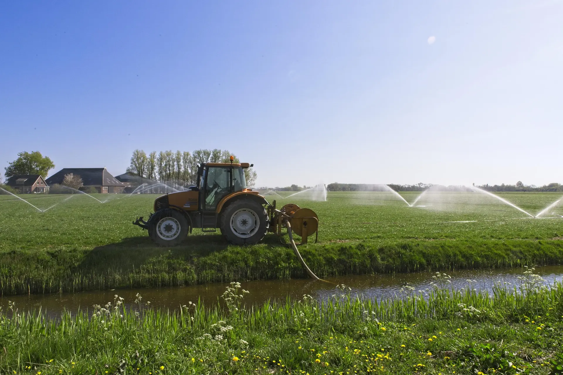 An orange tractor operates a multi-arm irrigation system, spraying water over a green field, with a water channel and wildflowers in the foreground.