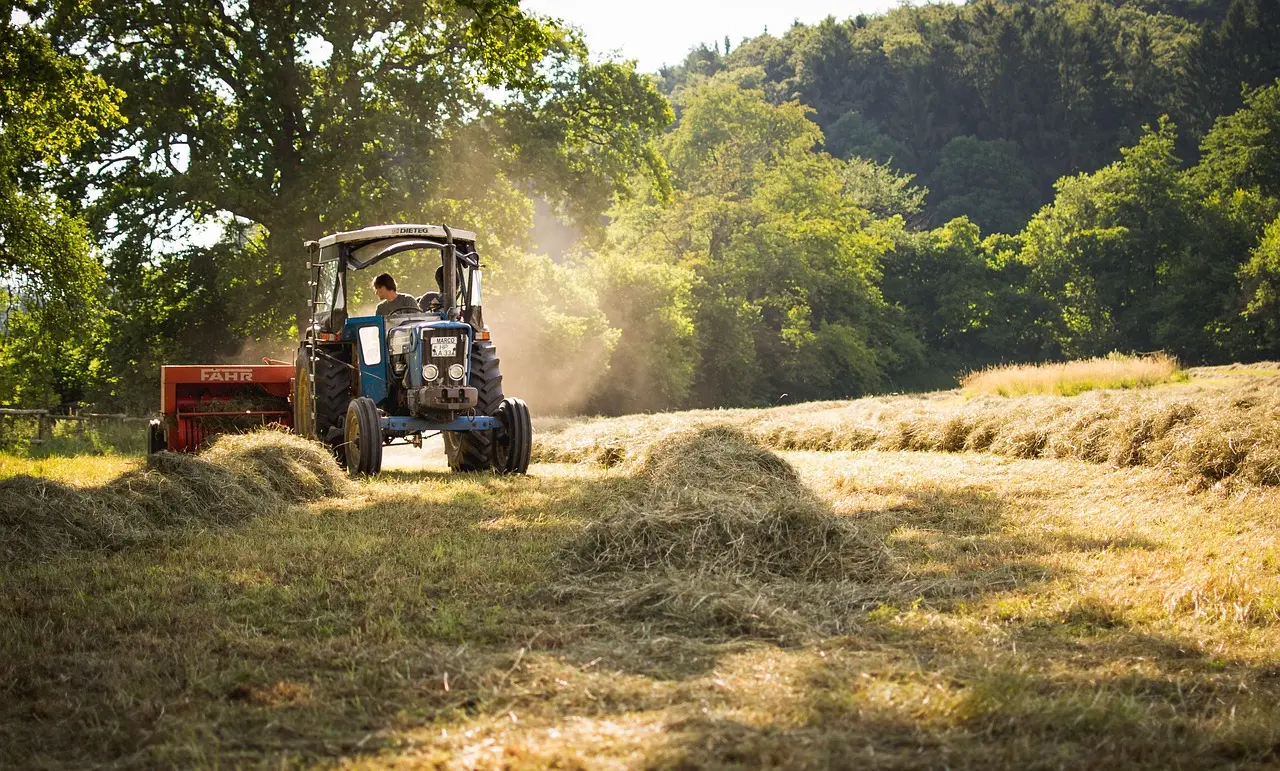 A blue FAHR tractor with an operator in the cab pulls a red implement through a field scattered with round hay bales, backed by dense green woodland and distant hills under a sunny sky.
