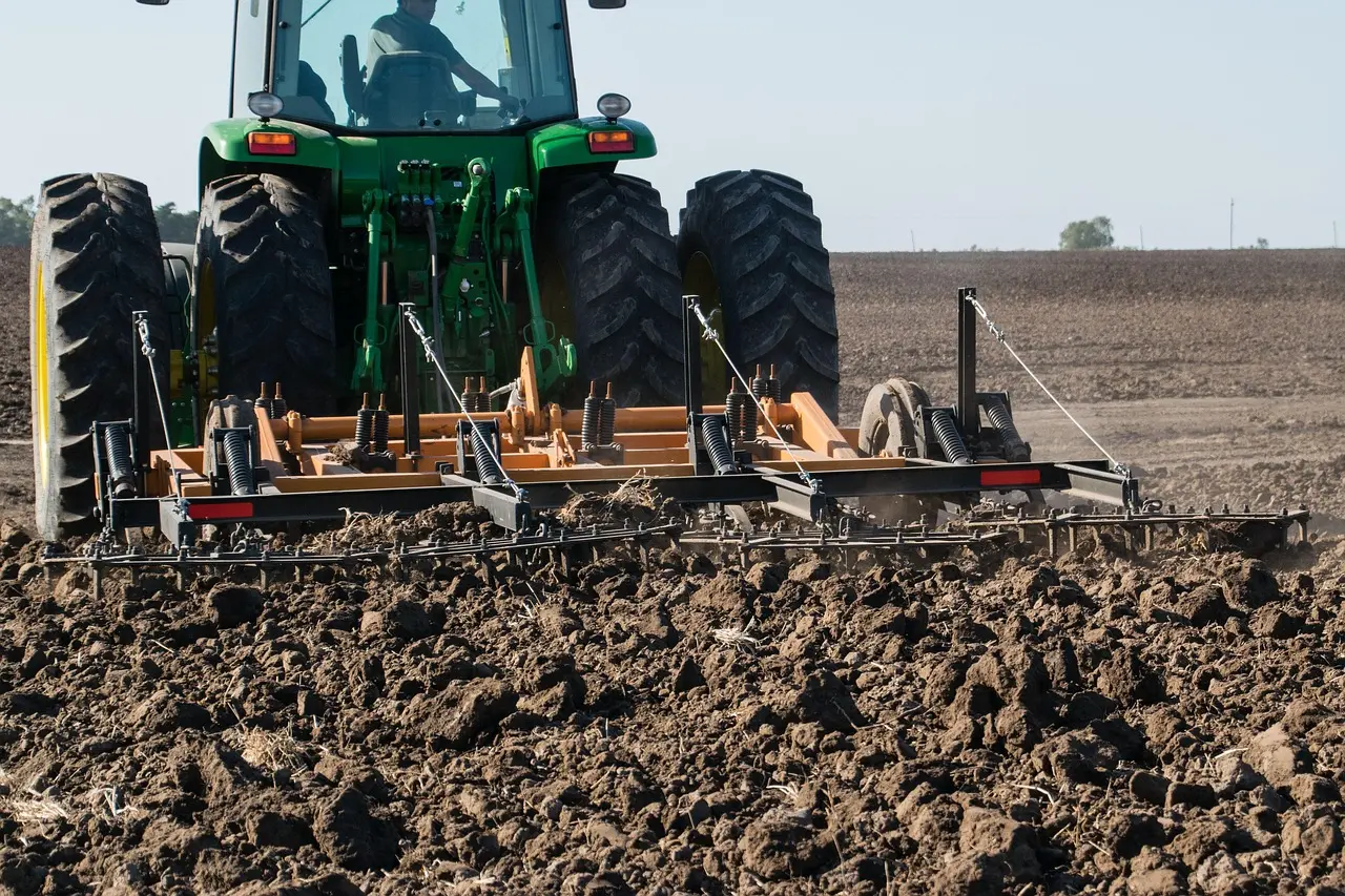 A green agricultural tractor equipped with a wide front-mounted tillage implement works through a field, breaking up and aerating the soil. The scene, dominated by earthy tones of green and brown, captures the tractor and implement in coordinated action, representing the fundamental process of soil preparation in farming.