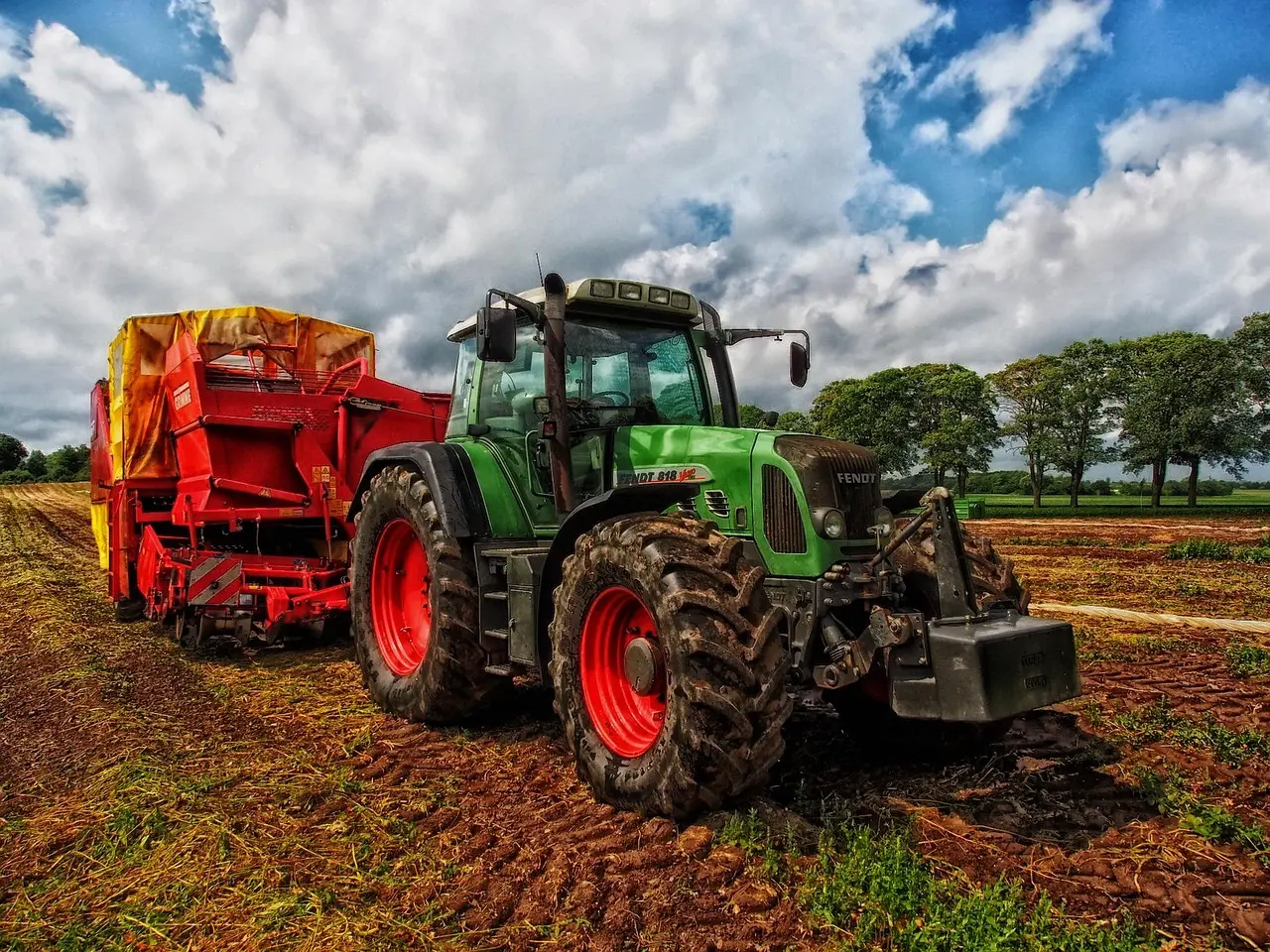 A robust green Fendt 818 Vario tractor with red front wheel hubs and muddy tires is parked on freshly worked soil. It is connected to a large red agricultural implement with an orange-yellow cover on top, all set against a dramatic cloudy sky.