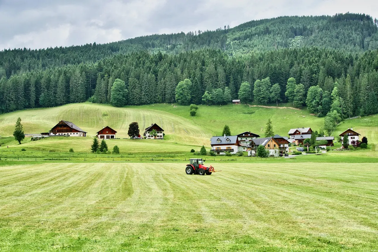 A peaceful and idyllic scene of alpine countryside: a red tractor sits on a lush green meadow leading to a traditional village and deep forests under a soft, cloudy sky, evoking a sense of tranquility and harmony with nature.