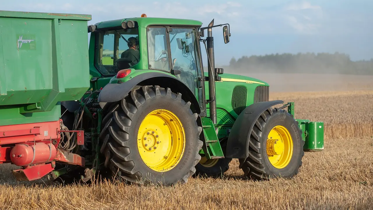 A powerful green John Deere tractor with distinctive yellow wheels hitched to a green trailer, parked in a vast golden wheat field under a soft blue sky, evoking the serenity of modern agricultural life.