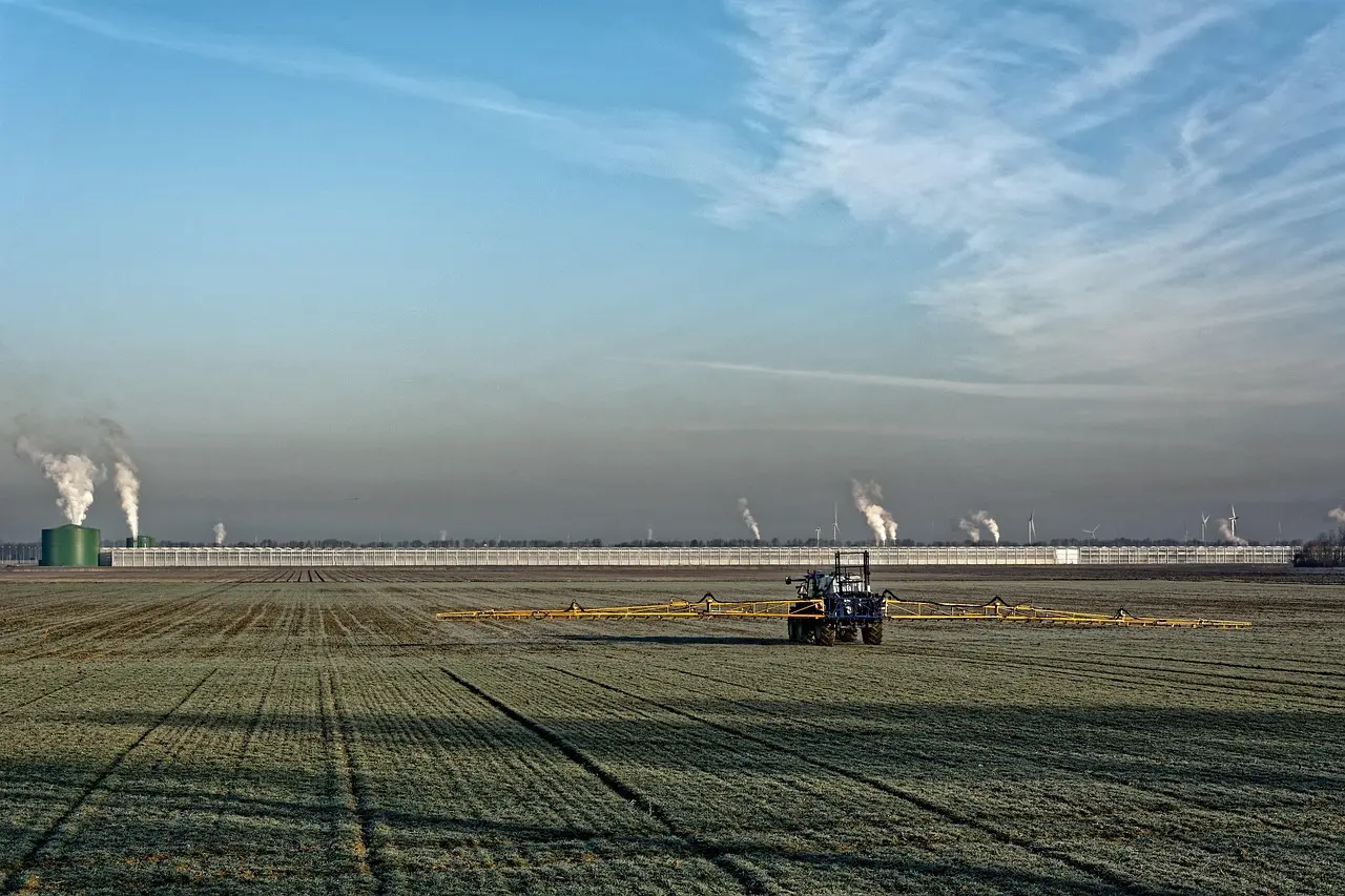 A scene of modern agriculture: a blue tractor farms a green field in the foreground, with the essential infrastructure of food production—industrial plants and wind turbines—visible in the distance.