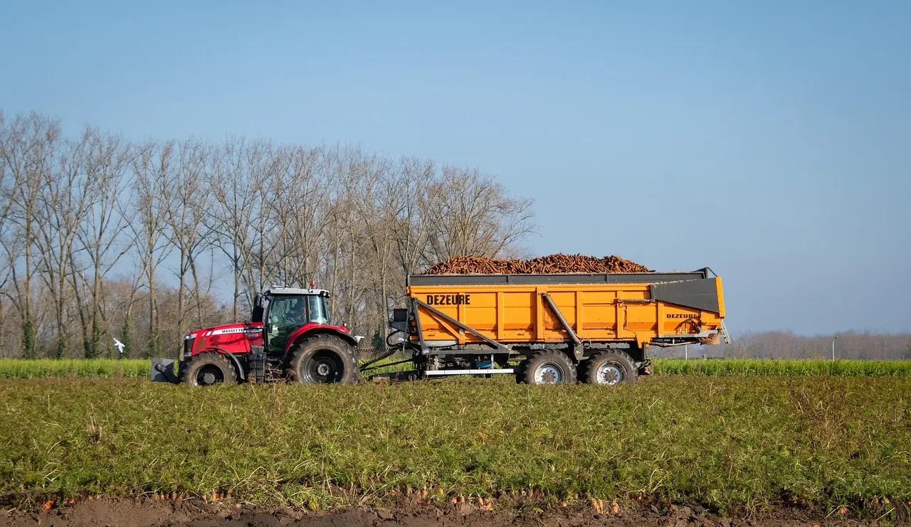 A red tractor parked in a green field, connected to a large orange trailer loaded with brown material (likely soil or harvested crop). The trailer bears the text "DE ZEURE" on its side, with bare trees and a clear blue sky in the background, depicting an agricultural work scene.