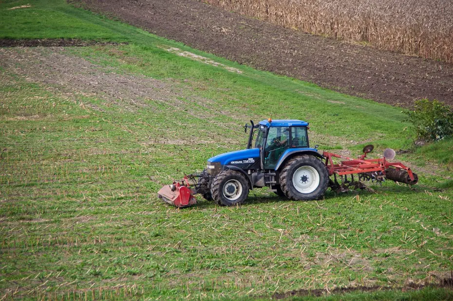 Modern agricultural technology in practice: A versatile blue tractor with implements pauses during field preparation, set against a backdrop that illustrates different stages of land management—from fresh tillage to mature crops—highlighting efficiency and the cycle of farm work.