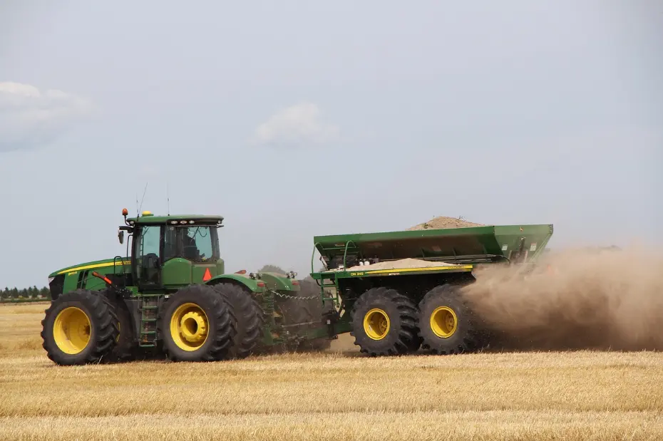 John Deere 9620RX tractor performing efficient material application in a wheat field. The large-scale equipment, with its powerful tires and spreader, embodies the strength and productivity of modern mechanized farming during a critical work period.
