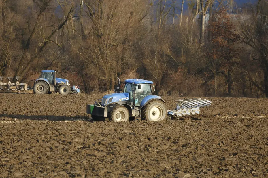Modern agricultural machinery in action: Blue tractors perform essential tillage in a damp field, preparing the soil for the coming season. The scene underscores the efficiency of mechanized farming even in cool, wet conditions.