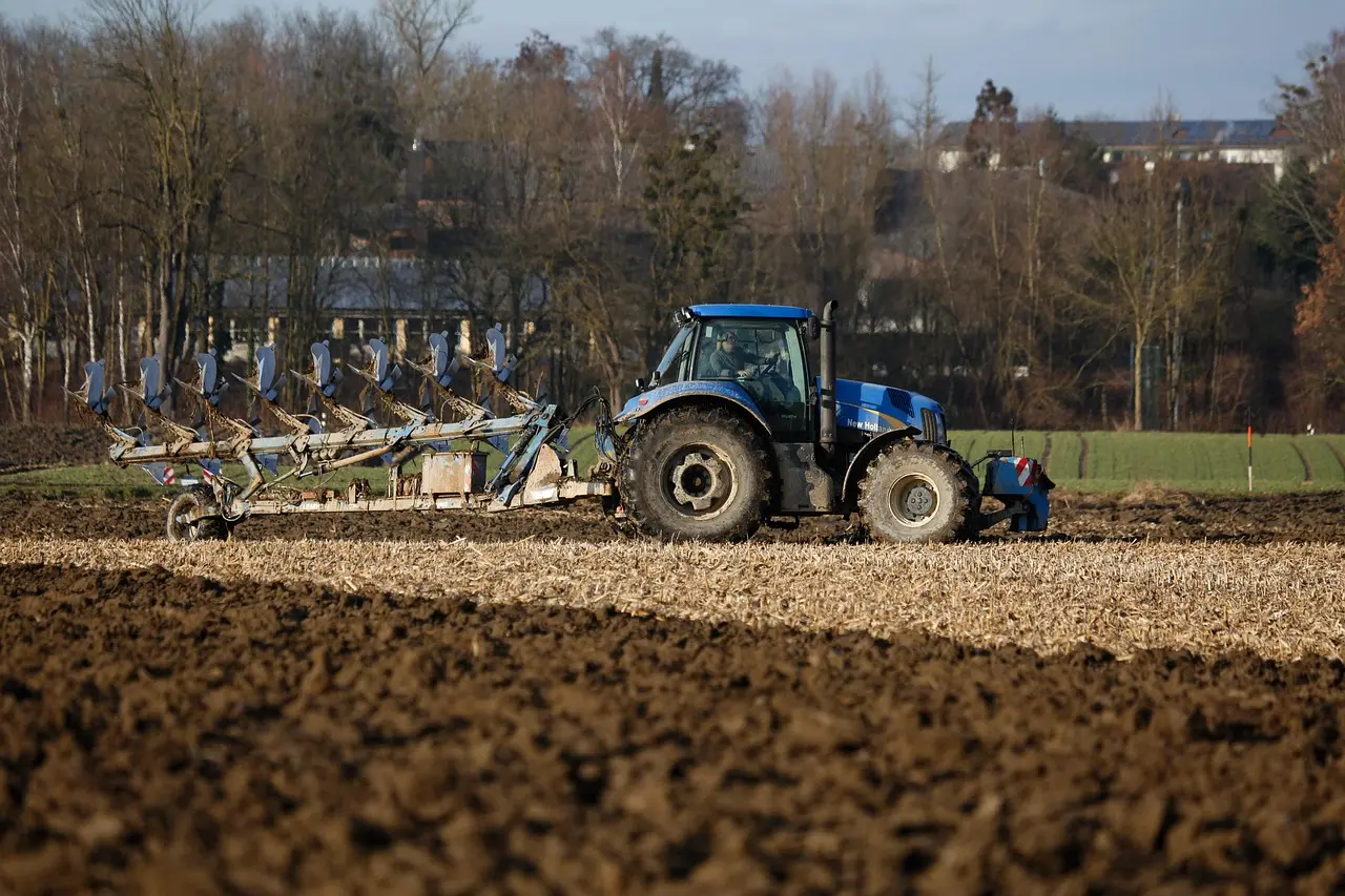 Efficient land preparation in action: a blue tractor and plow work the soil, showcasing the vital first step in the agricultural cycle. The scene emphasizes the scale and mechanical power required to prepare fields for planting.