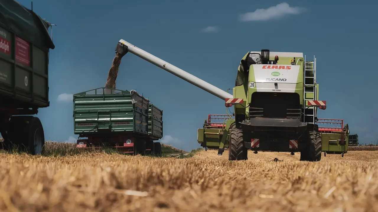 A CLAAS TUCANO 20 combine harvester unloads golden wheat into a green KRONE trailer via a long white chute during harvest. The powerful green machinery works in a vast, sunlit field of golden crops under a clear blue sky, capturing the efficiency and scale of modern mechanical agriculture.