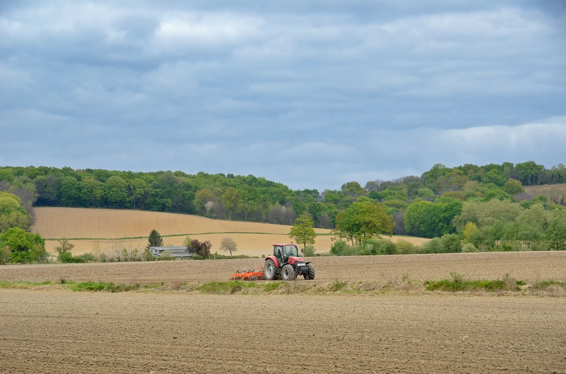 A red tractor tows an orange tillage implement (likely a disc harrow) across a vast, tilled field of uniform yellowish soil. A driver is visible in the cab. The expansive rural landscape features rolling hills covered in light brown farmland, bordered by lush green woodlands in the distance. The scene is set under a heavy, gray overcast sky, with the tractor positioned near the right-hand rule of thirds, creating a serene, layered, and vibrant depiction of spring or early summer fieldwork, rich in warm earth tones and greenery.